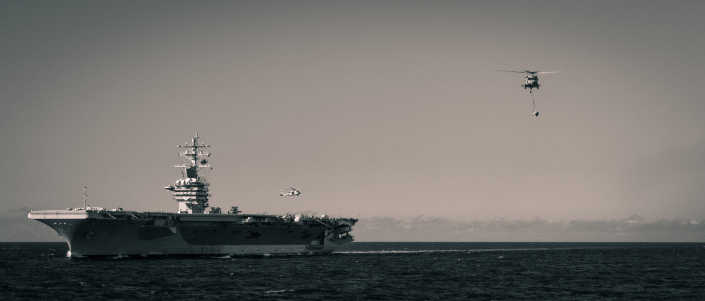B&W photo of an aircraft carrier with a helicopter hovering above it at sea.