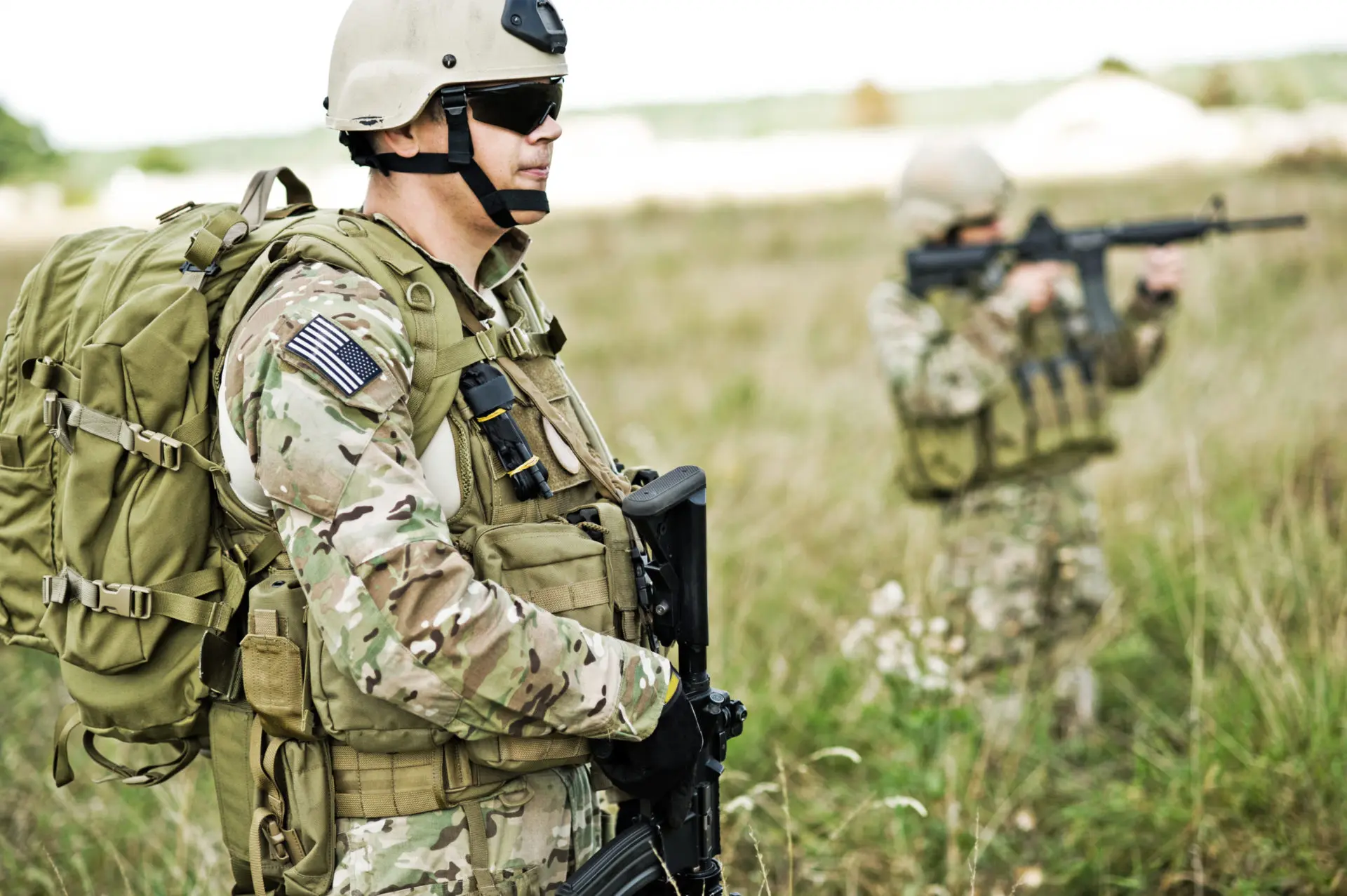 Two U.S. military personnel in camouflage, one with backpack and rifle, the other aiming a rifle.